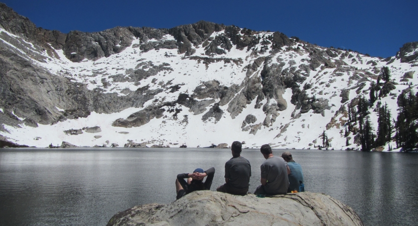 Three people sit on a large rock near a body of water. In the background, there is a snow-capped mountain. 